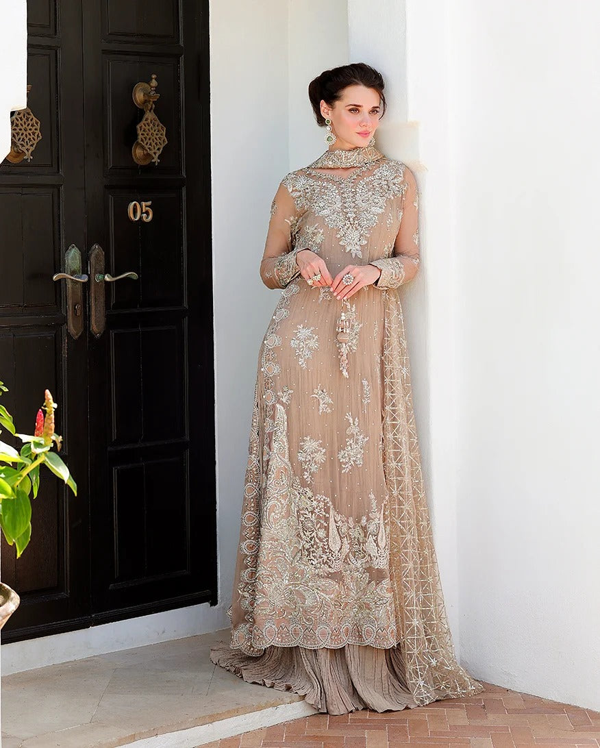 A woman models a floor-length beige embroidered gown with sheer sleeves and intricate floral embroidery, standing in front of a white wall and a dark wooden door.