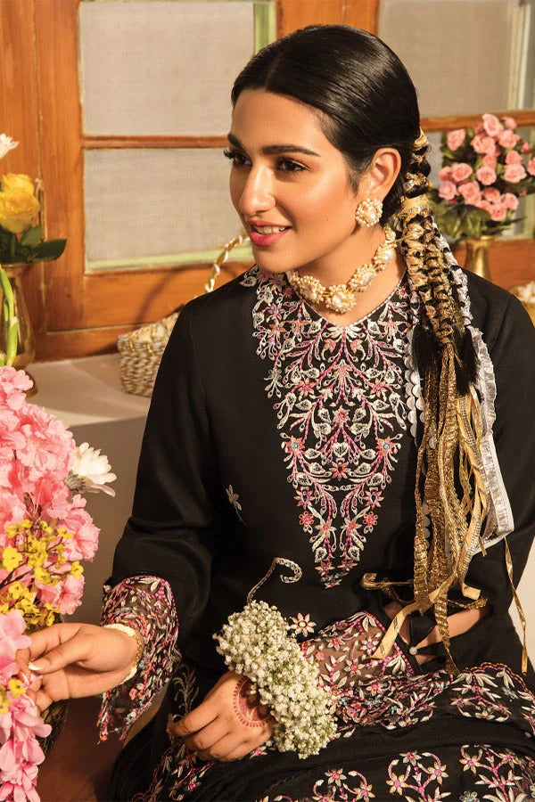 A woman wearing a black traditional outfit with ornate pink, white, and gold embroidery, holding a small bouquet of white flowers in a decorated indoor setting.