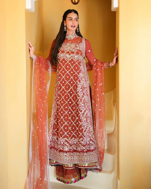 A woman models a traditional Indian salwar kameez outfit with an embroidered rust-orange and peach dupatta, set against a warm beige architectural background.