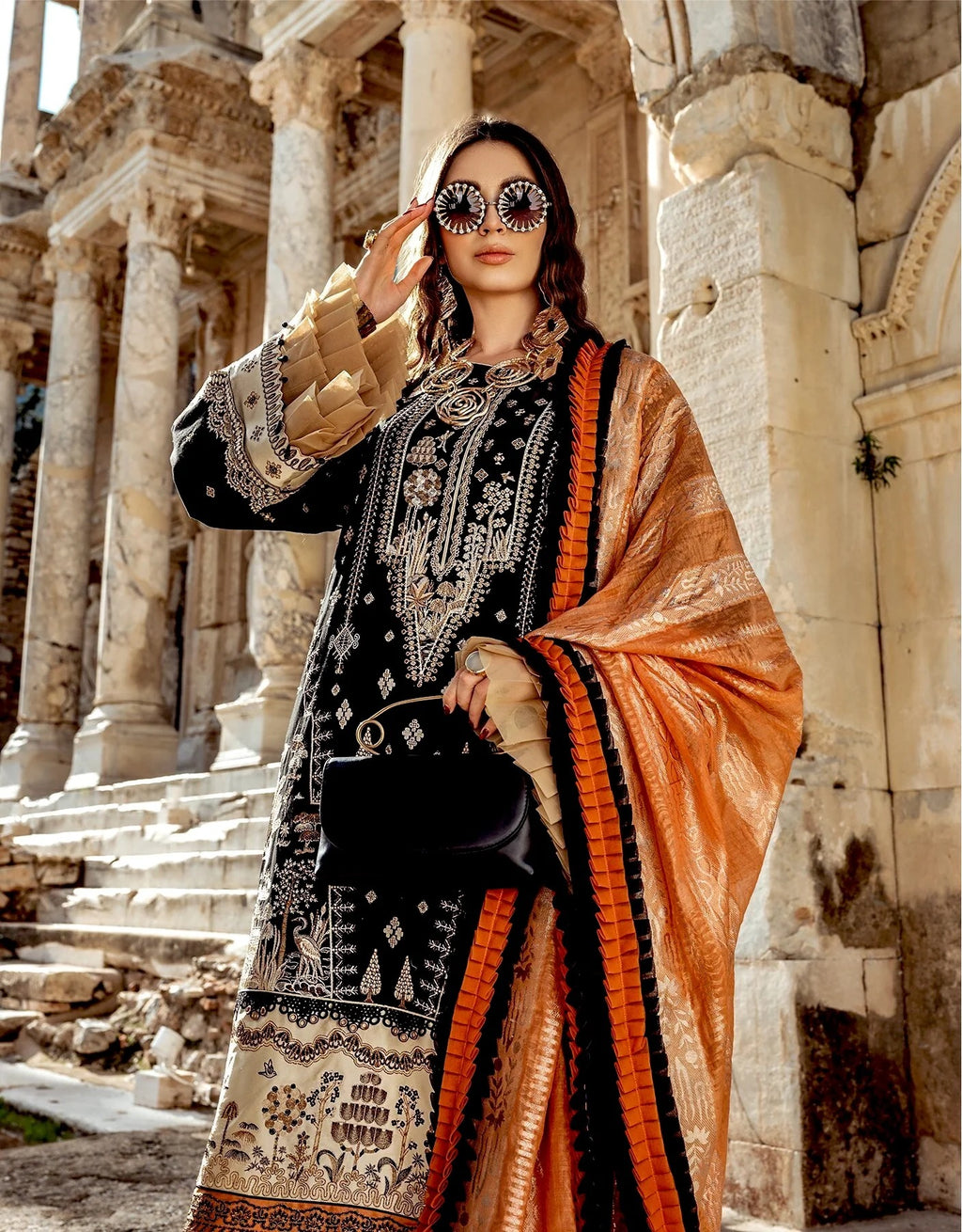 A woman stands in front of ancient stone columns wearing a black embroidered traditional outfit with ornate beige and silver patterns, a bright orange dupatta with black borders, and round sunglasses with mirrored lenses.