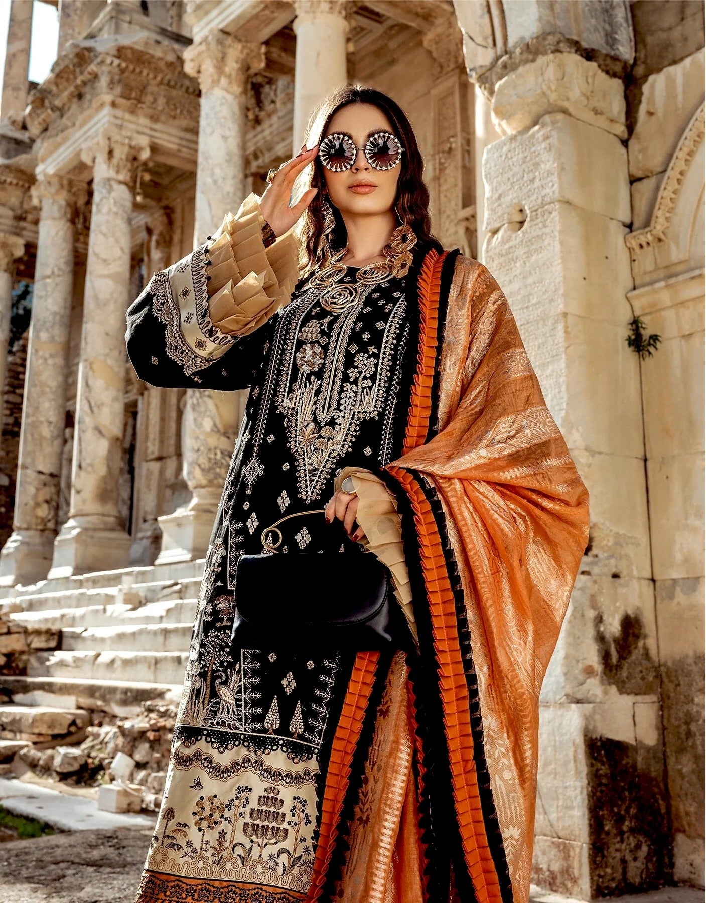 A woman stands in front of ancient stone columns wearing a black embroidered traditional outfit with ornate beige and silver patterns, a bright orange dupatta with black borders, and round sunglasses with mirrored lenses.