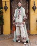 A woman stands in front of a rustic wooden door framed by yellow walls, wearing a traditional embroidered outfit. The outfit consists of an off-white kurta and wide-leg pants with colorful floral embroidery, a matching off-white dupatta with green borders, and a brown leather handbag.