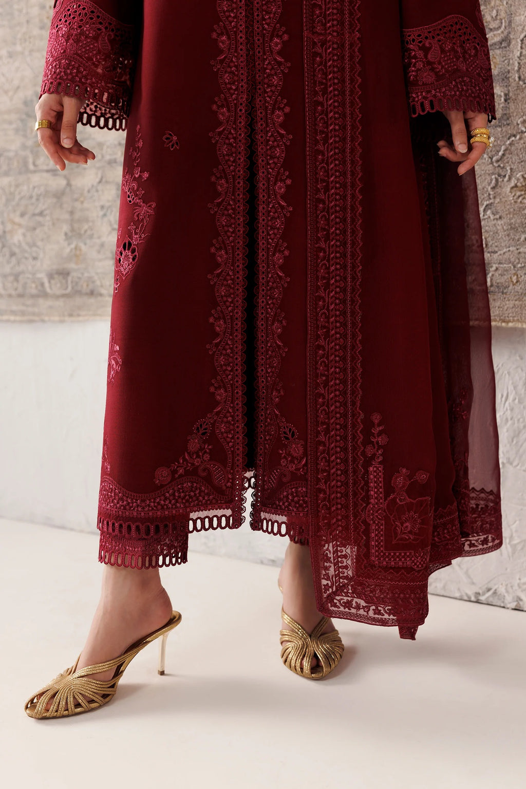 A person wearing a long burgundy embroidered dress or caftan with sheer panels and lace trim, paired with gold heeled sandals, standing against a light textured wall.