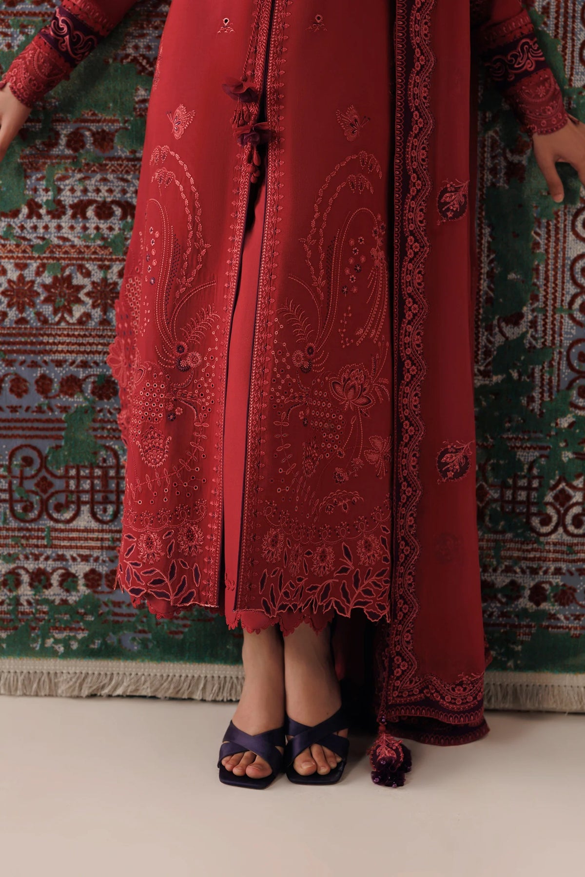 A person wearing a long red embroidered traditional outfit with matching sheer outer layer and dark purple sandals, standing against a patterned green rug.
