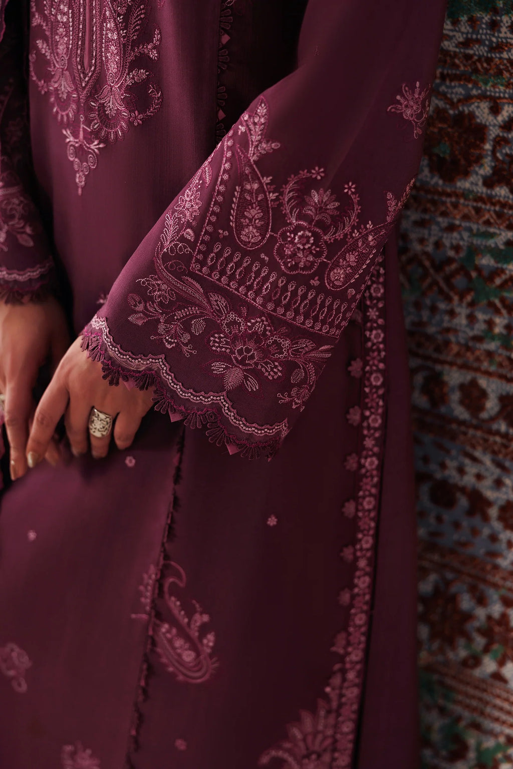 Close-up of a deep plum purple embroidered garment with intricate floral and paisley lace detailing on the sleeve cuffs and front panel, worn by a model whose hand with a ring is visible.