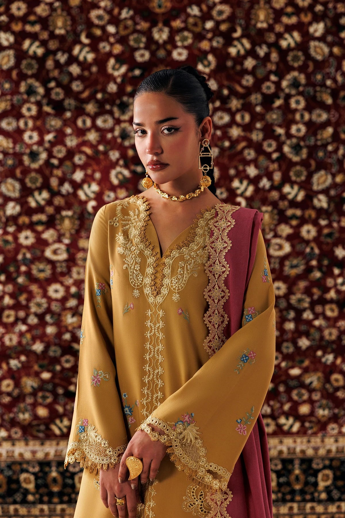 A woman wearing a traditional embroidered mustard yellow and deep pink kurta set with gold jewelry, standing in front of a patterned brown rug.