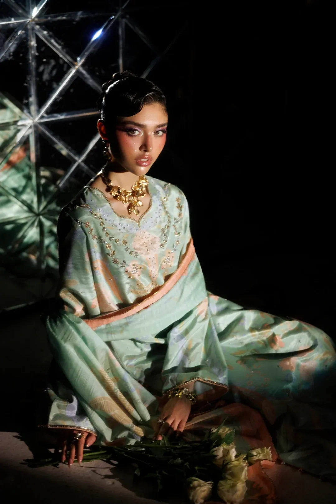 A model wearing a traditional Indian salwar kameez outfit in a light mint green color with gold embroidery, seated in a dark studio setting with dramatic lighting.
