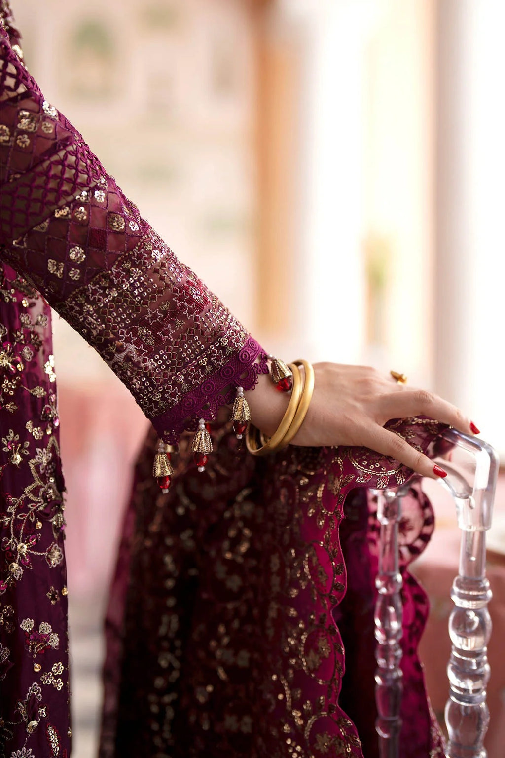 A close-up of a person's hand wearing gold bangles and holding a clear stand, dressed in an ornate deep magenta embroidered traditional outfit with gold detailing.