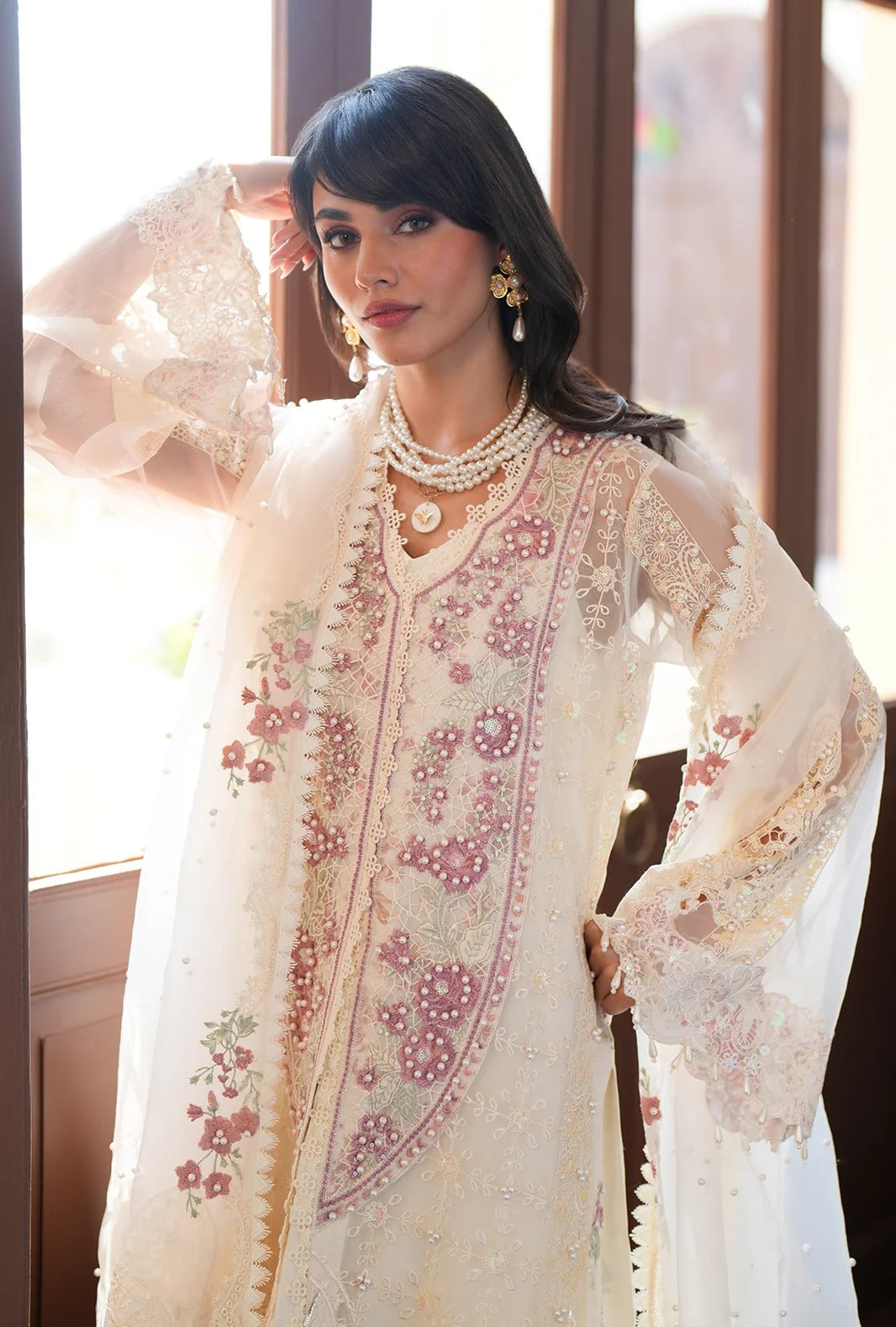 A woman models a traditional white and pink embroidered kurta set with sheer sleeves and floral embroidery, styled with layered pearl necklaces and earrings in a warm indoor setting.
