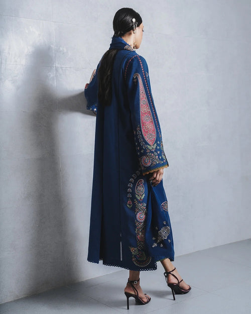A woman wearing a long navy blue embroidered kurta with intricate red, gold, and white paisley patterns, standing against a gray concrete wall.
