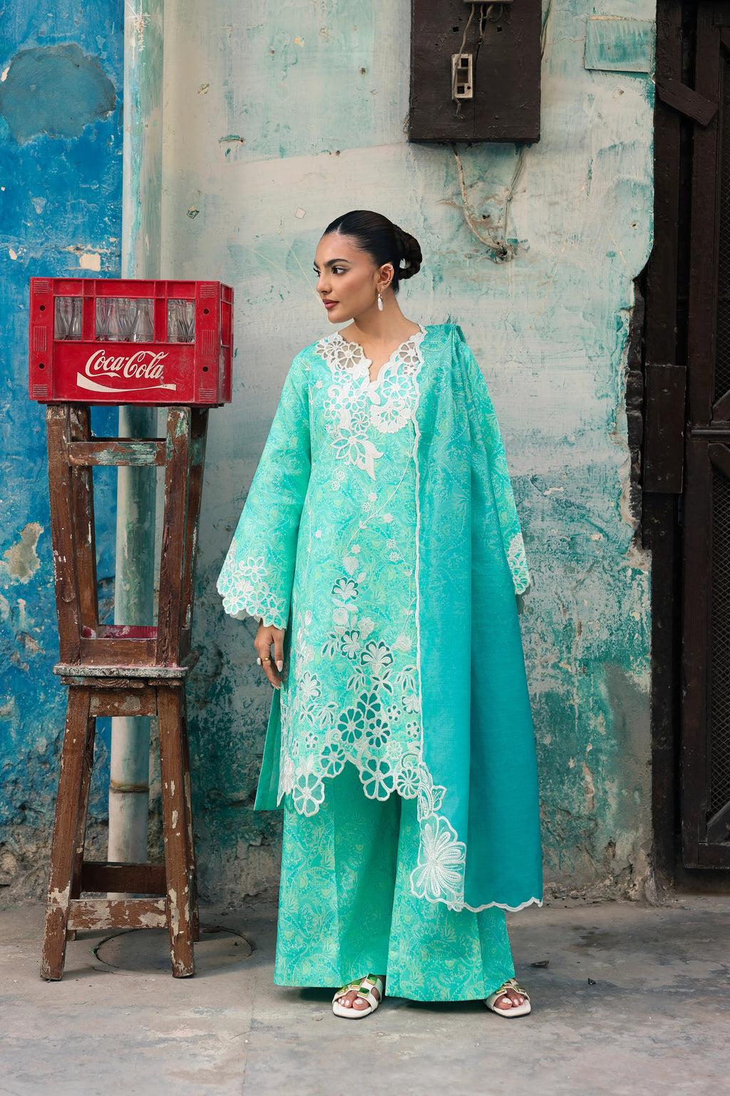 A woman models a mint green traditional outfit with white floral embroidery, standing in front of a weathered blue wall next to a red Coca-Cola crate on a wooden stand.
