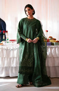 A woman models a dark green traditional outfit with gold embroidery, wide-leg pants, and a matching sheer dupatta in an indoor event setting.