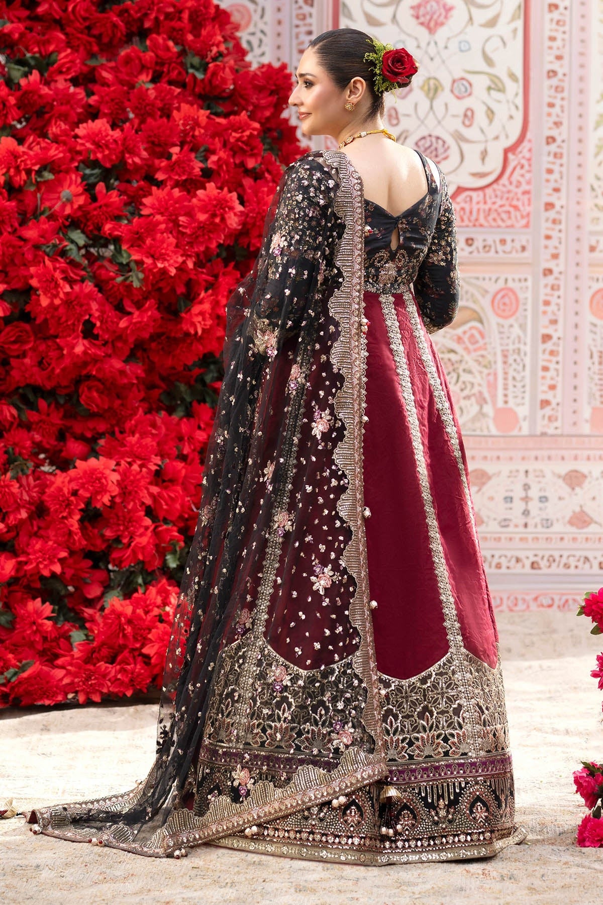 A woman wearing an ornate traditional Indian lehenga choli dress, photographed from the back. The dress features a deep maroon skirt with heavy gold and silver embroidery, a black embroidered choli, and a matching sheer black dupatta draped over her shoulder.