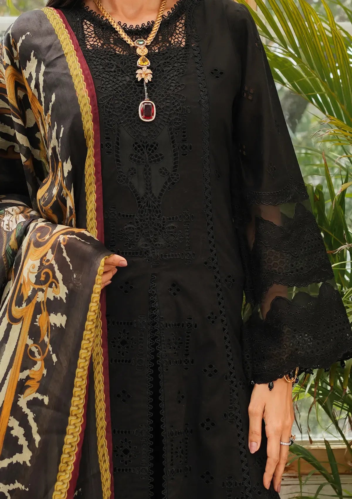 A close-up of a person wearing a black embroidered kurta with sheer sleeves and a patterned dupatta, styled with a statement necklace in front of a green plant background.