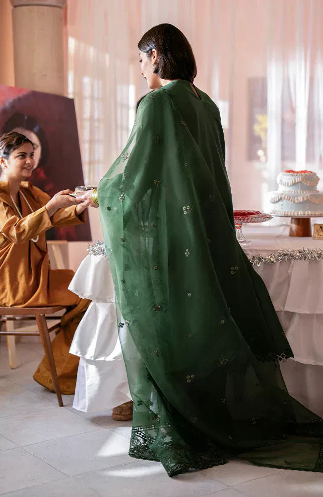A woman in a traditional dark green embroidered shawl and matching outfit stands with her back to the camera in a softly lit indoor setting, while another woman in a mustard outfit sits nearby.