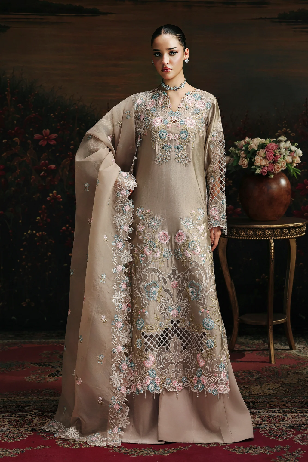 A woman models an elegant beige traditional outfit with intricate multicolor floral embroidery, a matching dupatta, and a long skirt, set against a dark, ornate background with a floral arrangement and a side table.