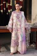 A woman stands indoors wearing a long cream-colored embroidered kurta with pink, purple, and blue floral motifs, paired with matching wide-leg pants and a sheer pastel dupatta draped over one shoulder. The setting includes a rustic wooden table, hanging garlands, and textured walls.