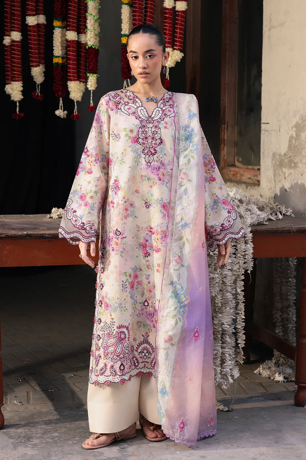 A woman stands indoors wearing a long cream-colored embroidered kurta with pink, purple, and blue floral motifs, paired with matching wide-leg pants and a sheer pastel dupatta draped over one shoulder. The setting includes a rustic wooden table, hanging garlands, and textured walls.