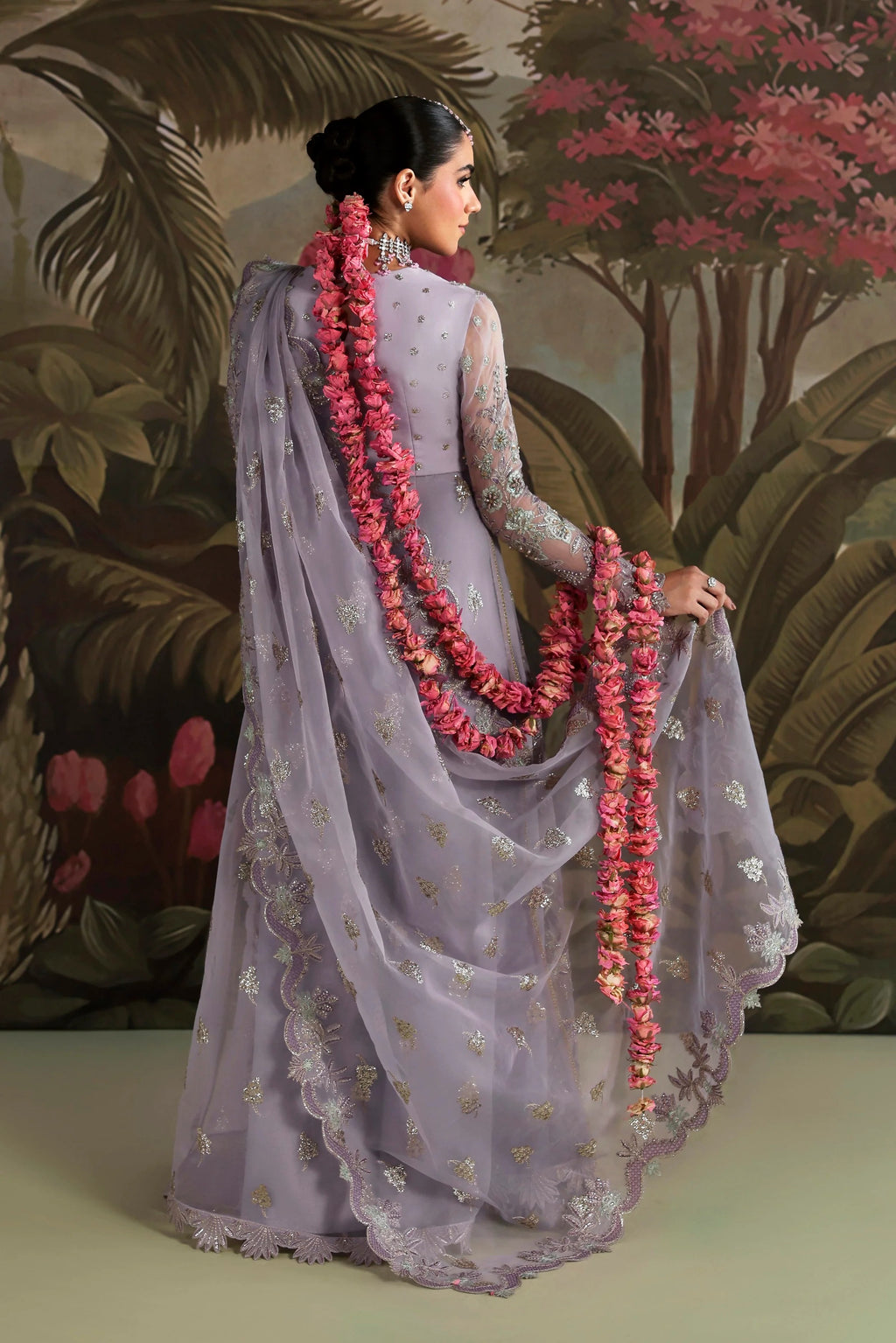 A woman wearing a lavender-purple embroidered traditional outfit with a matching sheer dupatta and long floral garlands, posed in front of a tropical forest backdrop.