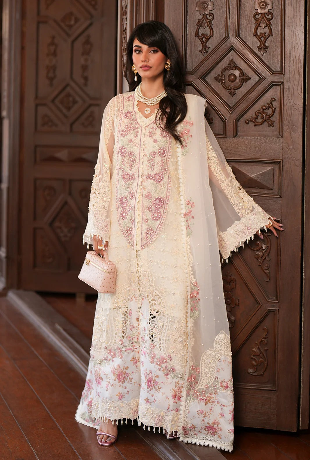A woman models a traditional cream-colored embroidered Anarkali gown with pink floral patterns and a matching sheer dupatta, standing in front of an ornate dark wood door.