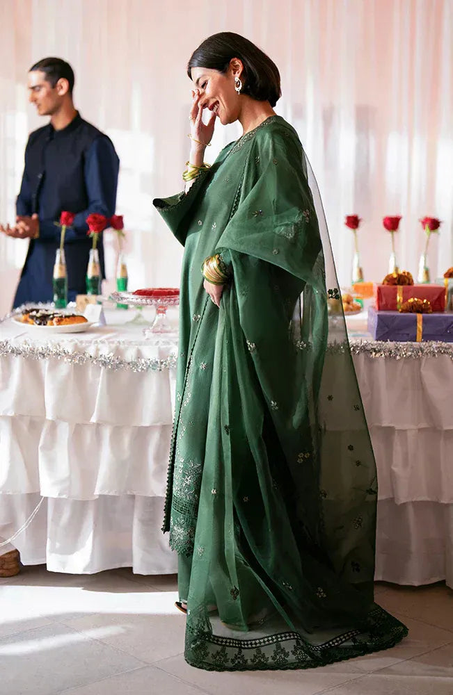 A woman wearing a dark green embroidered traditional outfit with a sheer dupatta stands in a decorated indoor event space with tables, gifts, and a man blurred in the background.