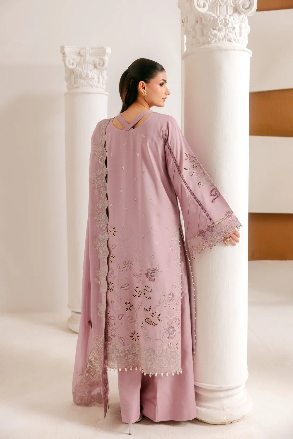 A woman wearing a dusty pink traditional outfit with floral embroidery and gold mirror work, shown from the back as she stands between two white columns in a bright indoor setting.