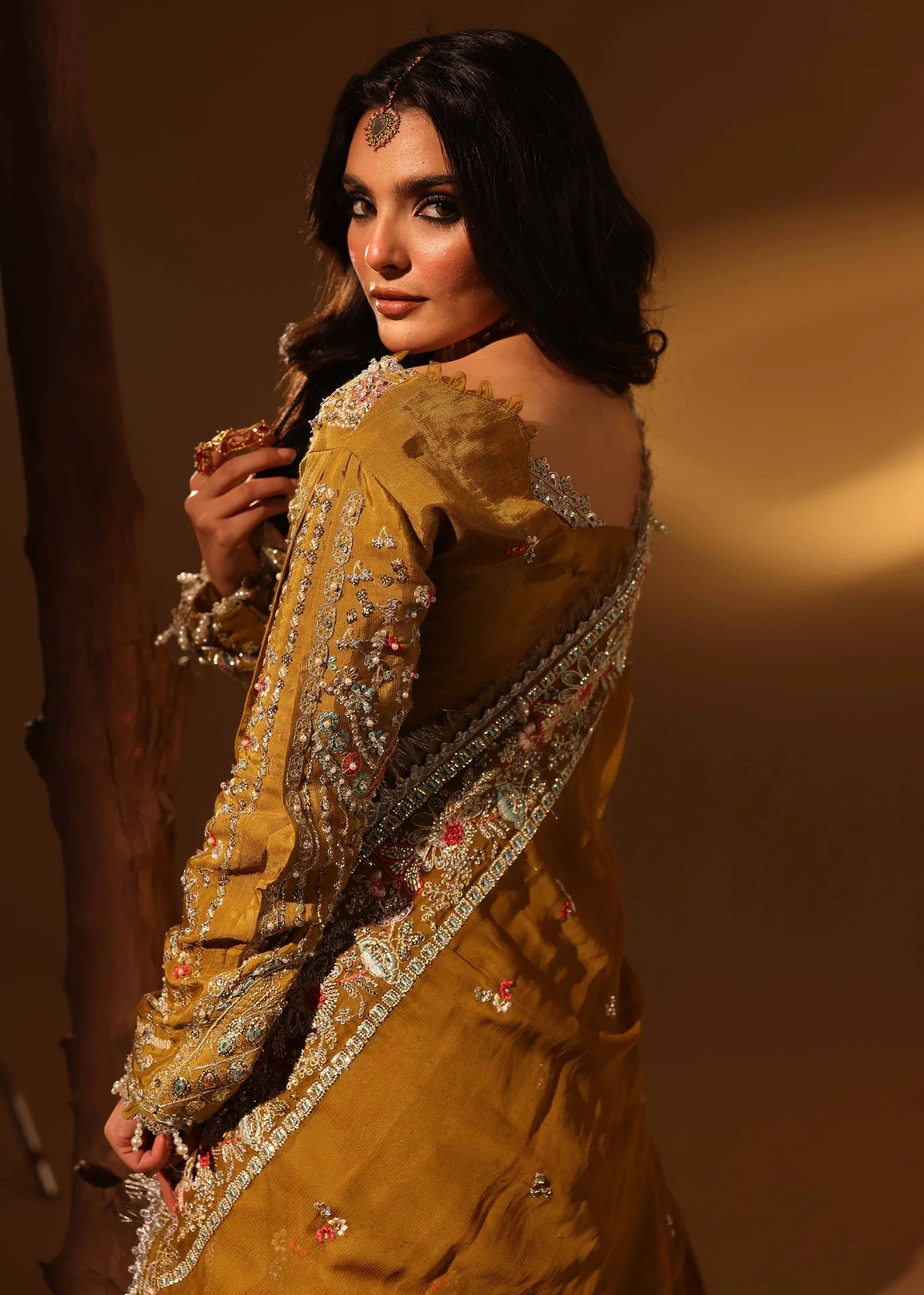 Woman in a mustard yellow traditional outfit with intricate detailing against a warm brown background