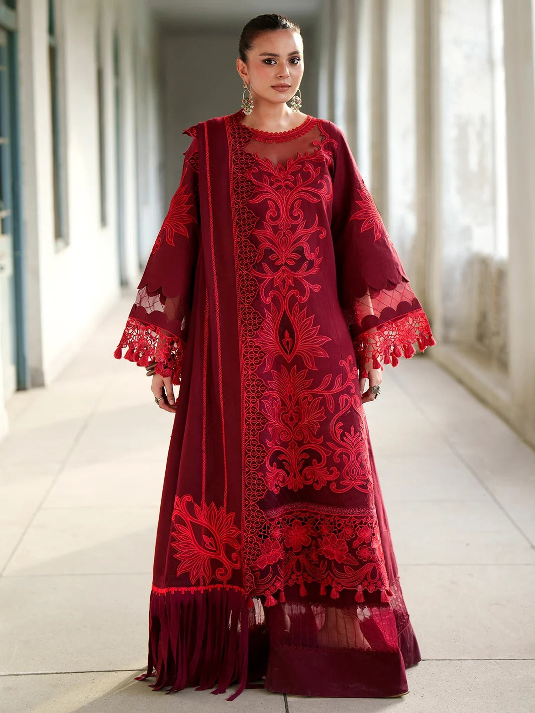A woman models a deep red embroidered traditional dress with matching dupatta in a bright, sunlit corridor.