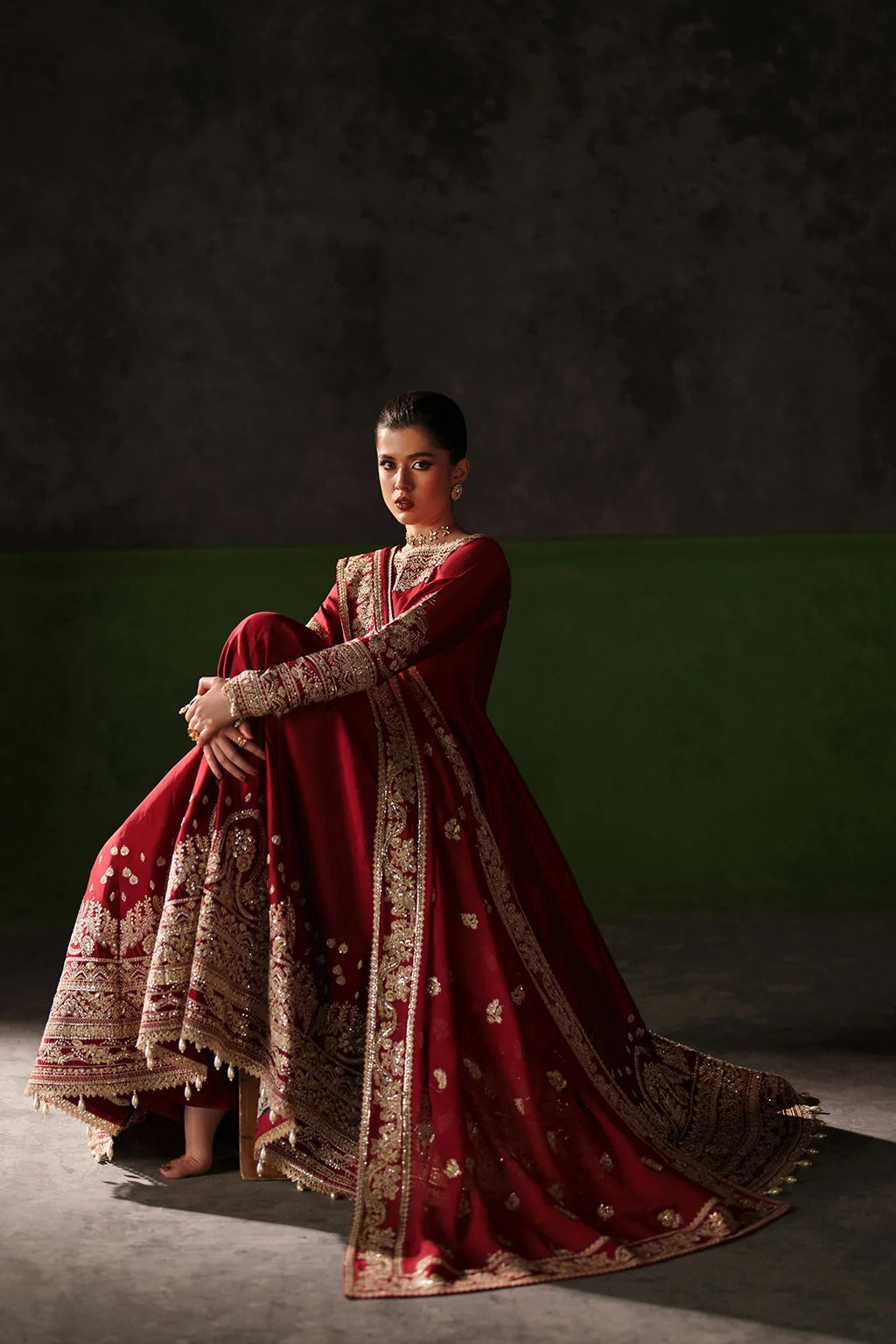 A woman models a traditional red and gold embroidered Anarkali gown against a dark studio backdrop with a green lower panel.