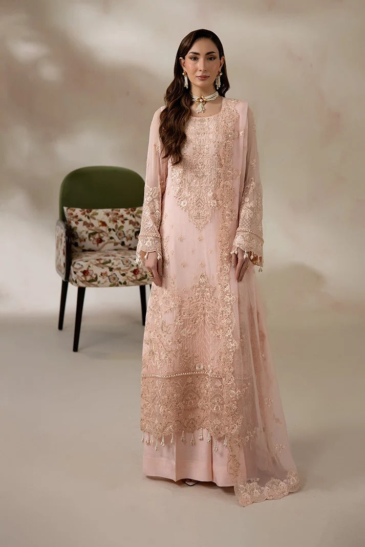 A woman models a long-sleeved light pink traditional gown with intricate gold embroidery and a matching sheer dupatta, set against a warm beige studio backdrop with a green accent chair.