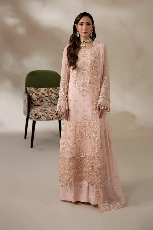 A woman models a long-sleeved light pink traditional gown with intricate gold embroidery and a matching sheer dupatta, set against a warm beige studio backdrop with a green accent chair.