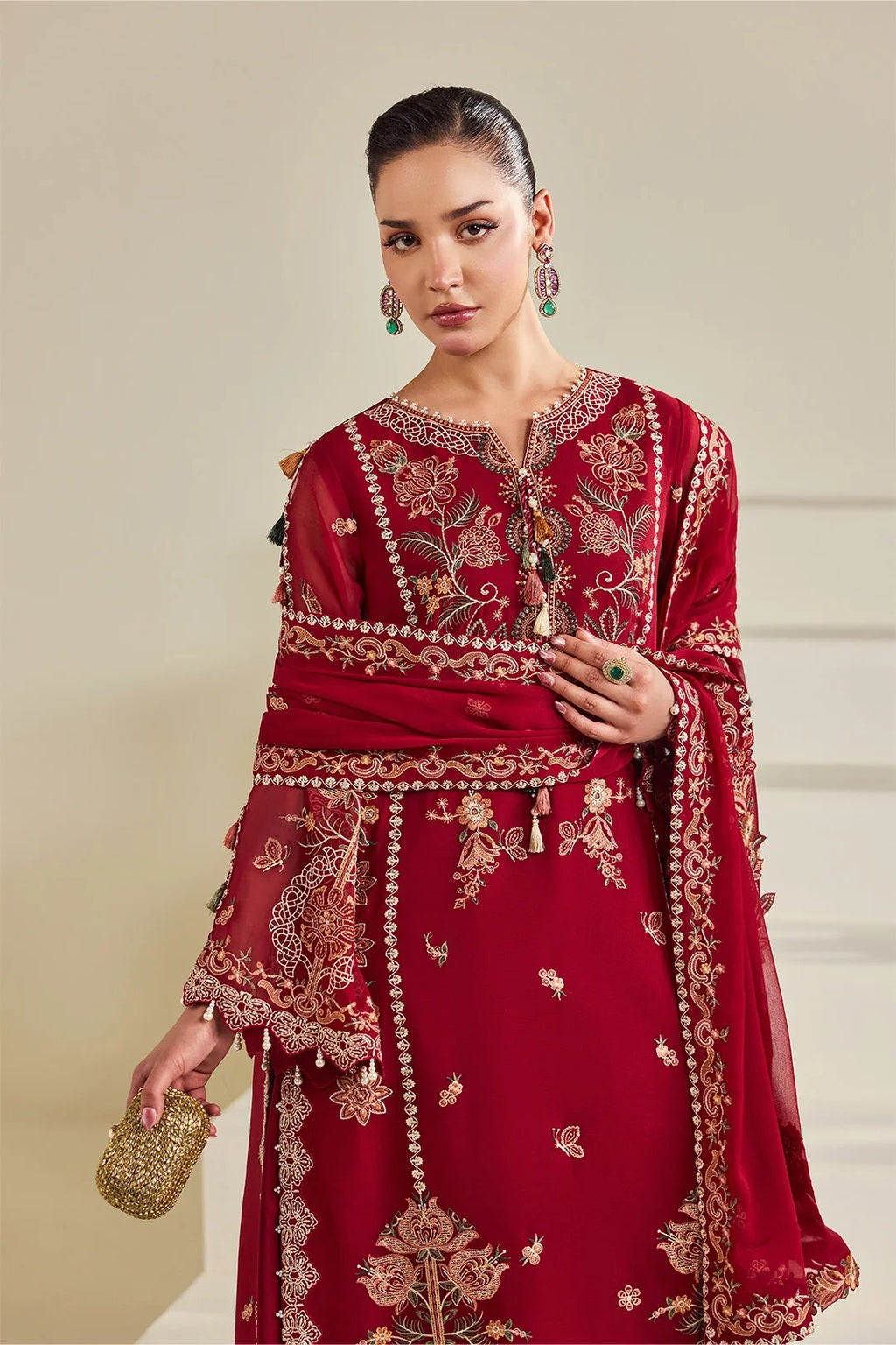 A woman models a traditional deep red embroidered outfit with gold and beige floral detailing, a matching dupatta, and a small gold clutch against a neutral beige background.