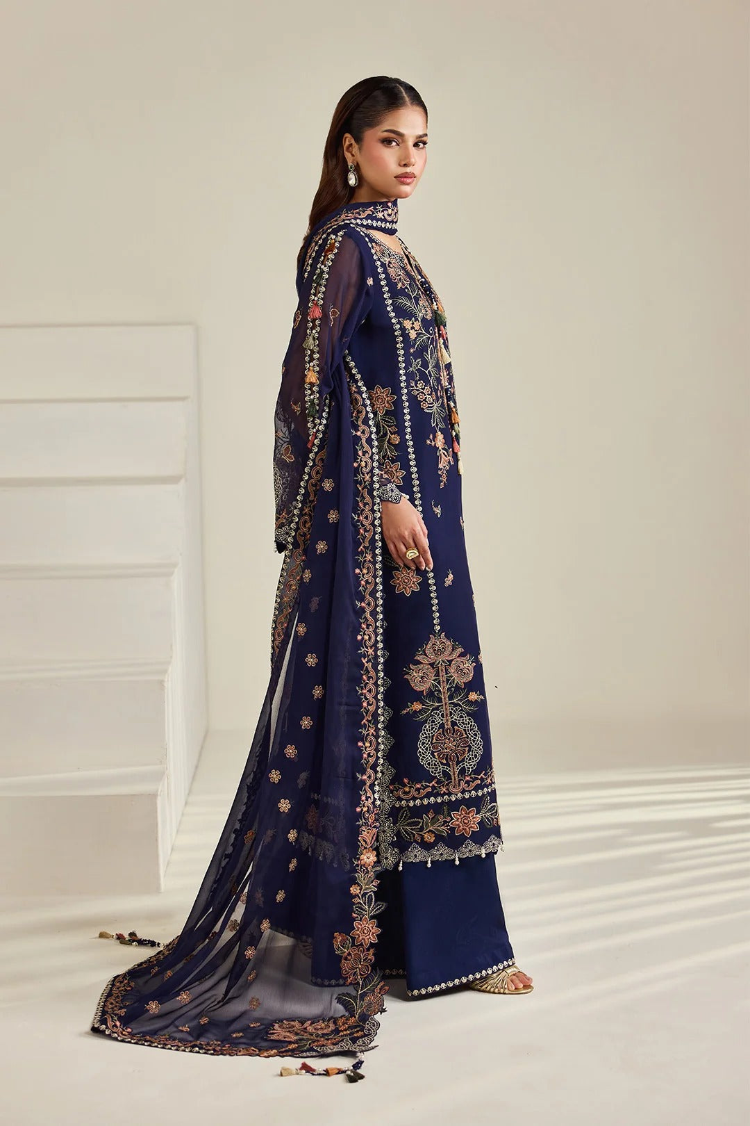 A woman modeling a navy blue traditional outfit with intricate gold and beige embroidery, set against a minimalist cream-colored studio background.