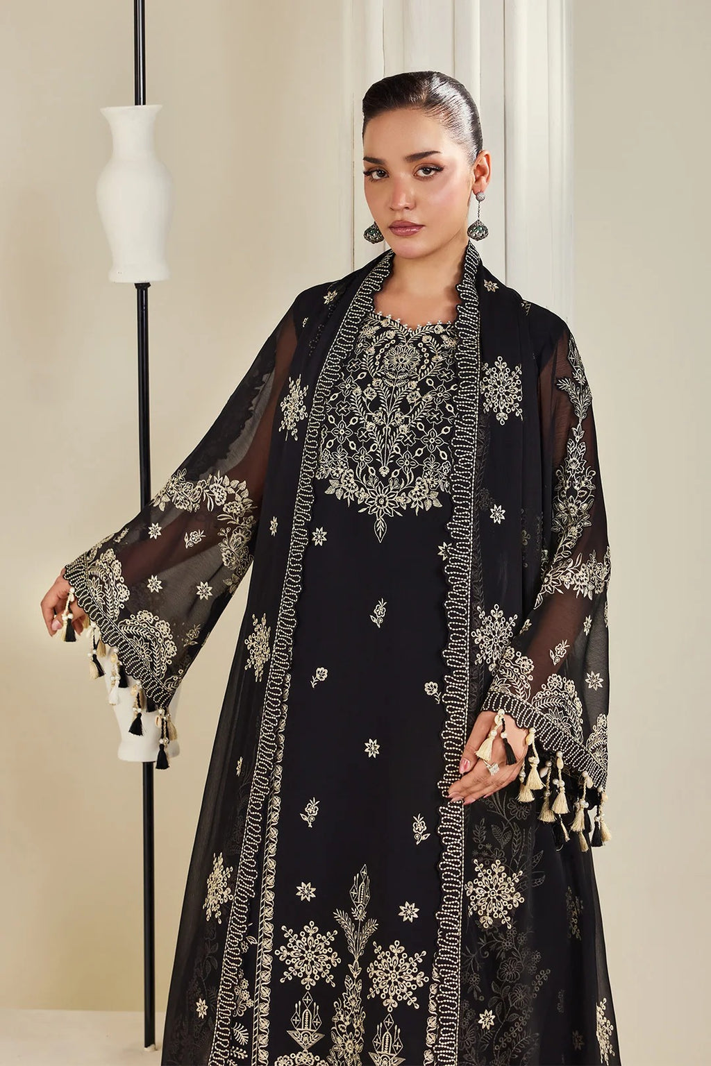 A woman models a black embroidered traditional outfit with sheer sleeves and gold-toned embellishments, standing indoors against a light neutral wall beside a white vase on a stand.