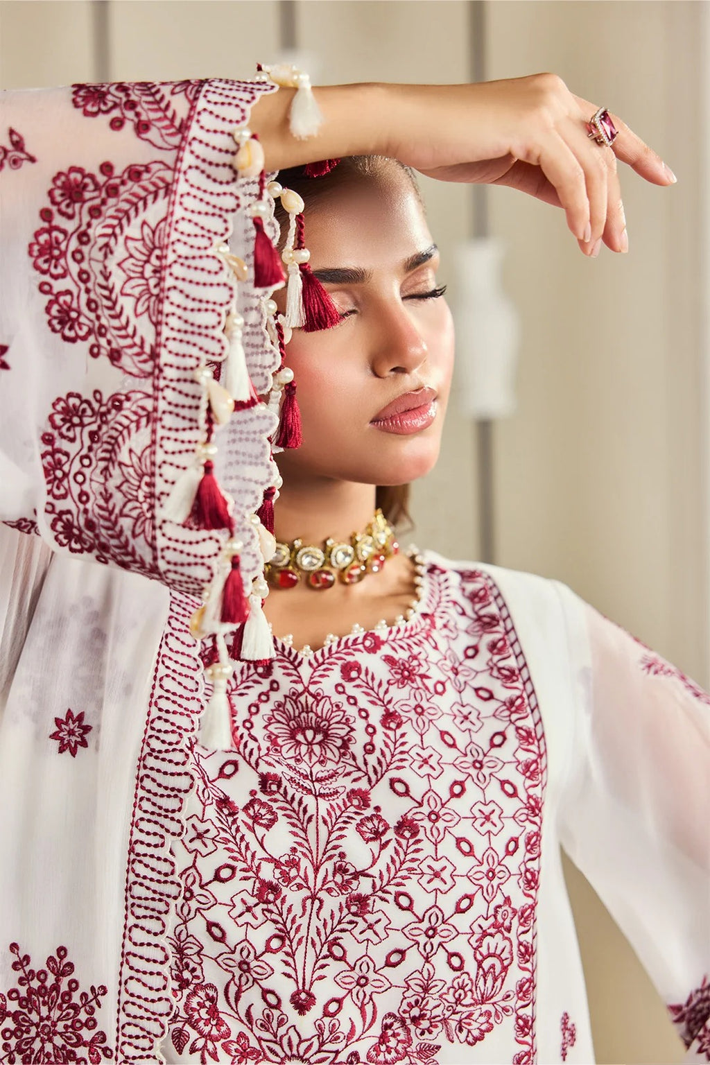 A woman wearing a white traditional embroidered outfit with deep red floral embroidery and tassel details on the sleeve, paired with a matching red-and-white dupatta and gold jewelry.