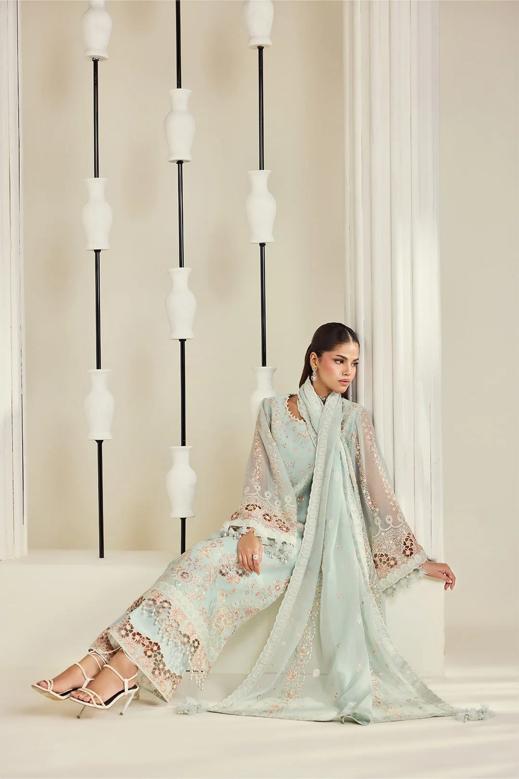 A woman models a light mint green ethnic three-piece suit with intricate gold embroidery and a matching sheer dupatta, posed in a minimalist cream-colored room with decorative vertical light fixtures.