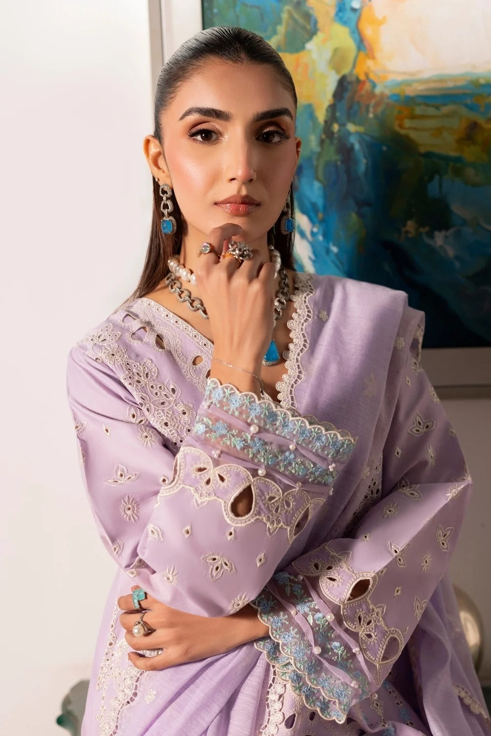 Woman wearing a lavender traditional outfit with intricate designs, standing in front of a colorful abstract painting.