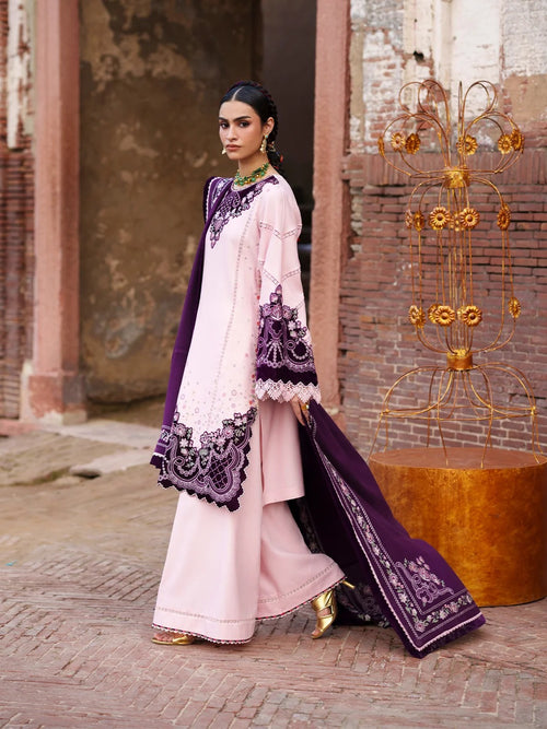 A woman models a traditional light pink three-piece salwar kameez suit with deep purple embroidery and a matching purple dupatta, set against a rustic brick and stone background.