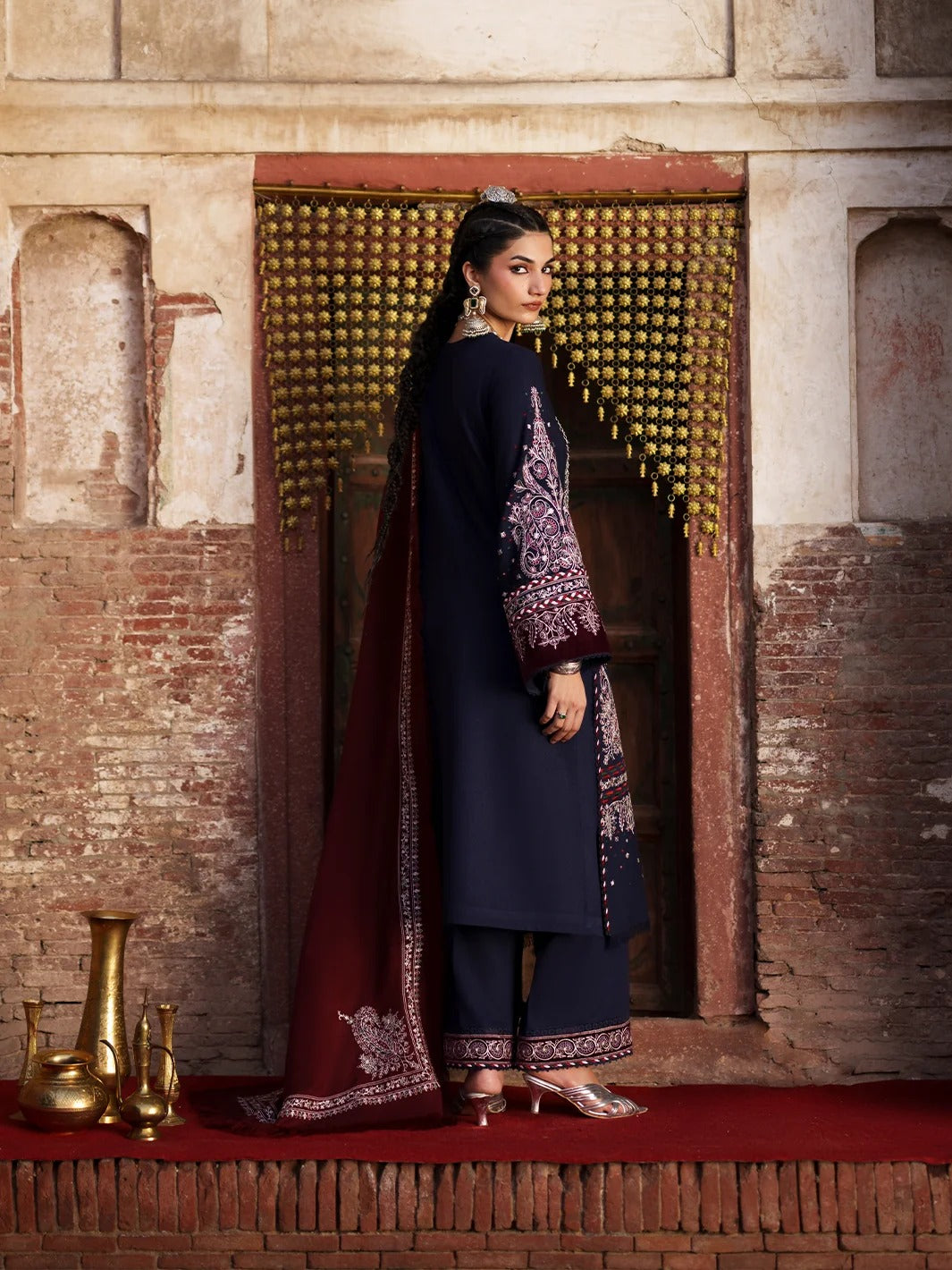 A woman models a traditional Indian outfit consisting of a navy blue kurta with purple and gold embroidery, matching navy pants, and a deep maroon dupatta with gold border detailing, set against a rustic brick and stone wall background.