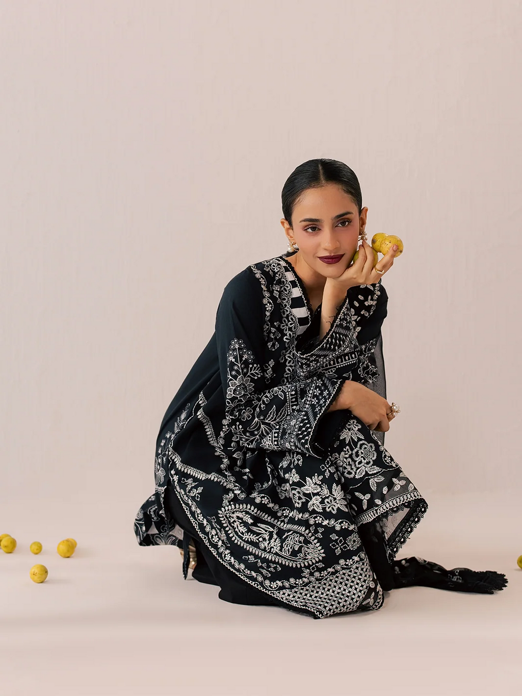 A woman models a black and white printed traditional outfit with a matching dupatta, posing on a light beige studio background with scattered yellow fruits.