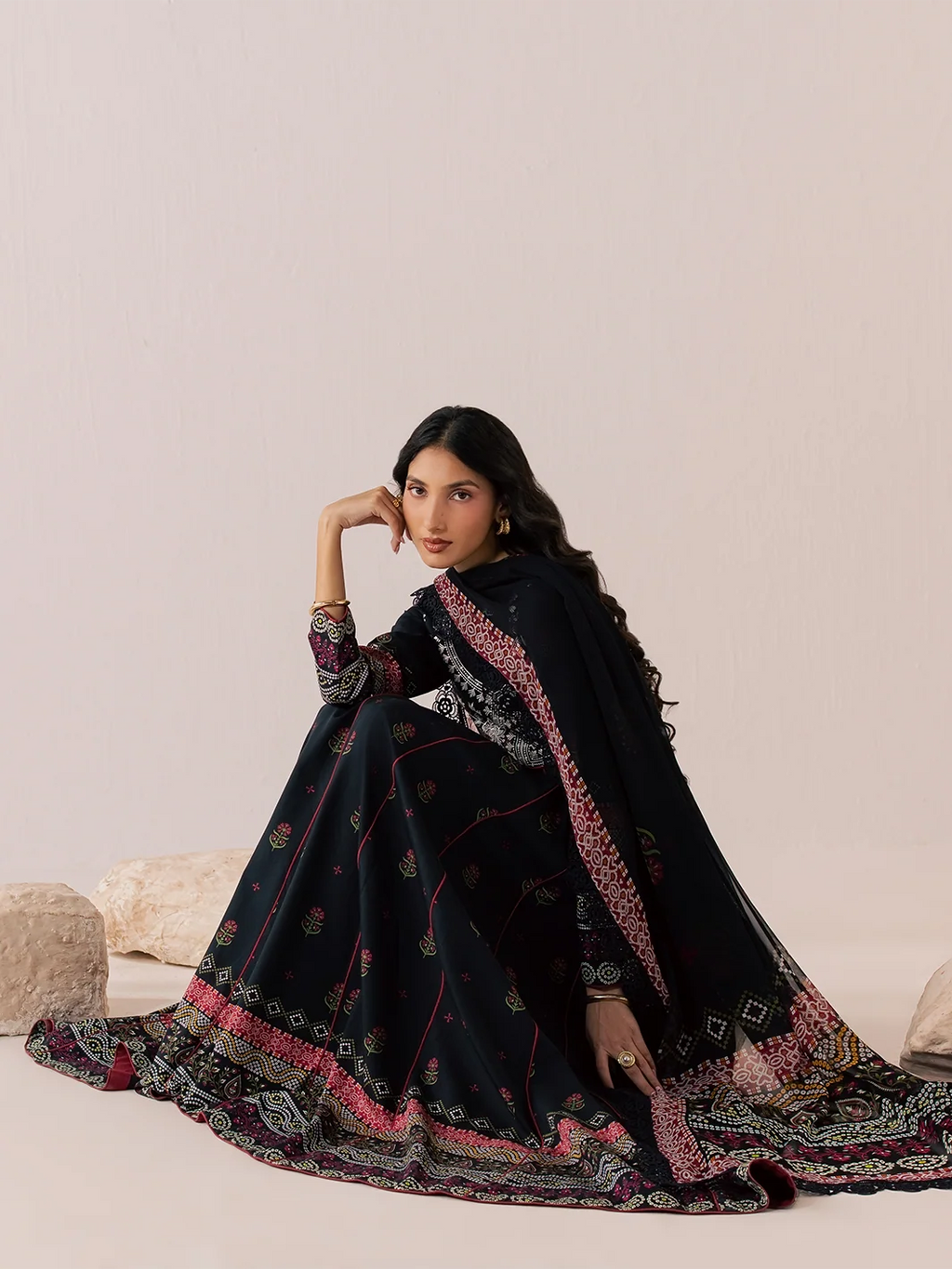 A woman models a black three-piece traditional outfit with intricate pink, red, and gold floral prints and borders, posed against a neutral beige background with decorative stones.