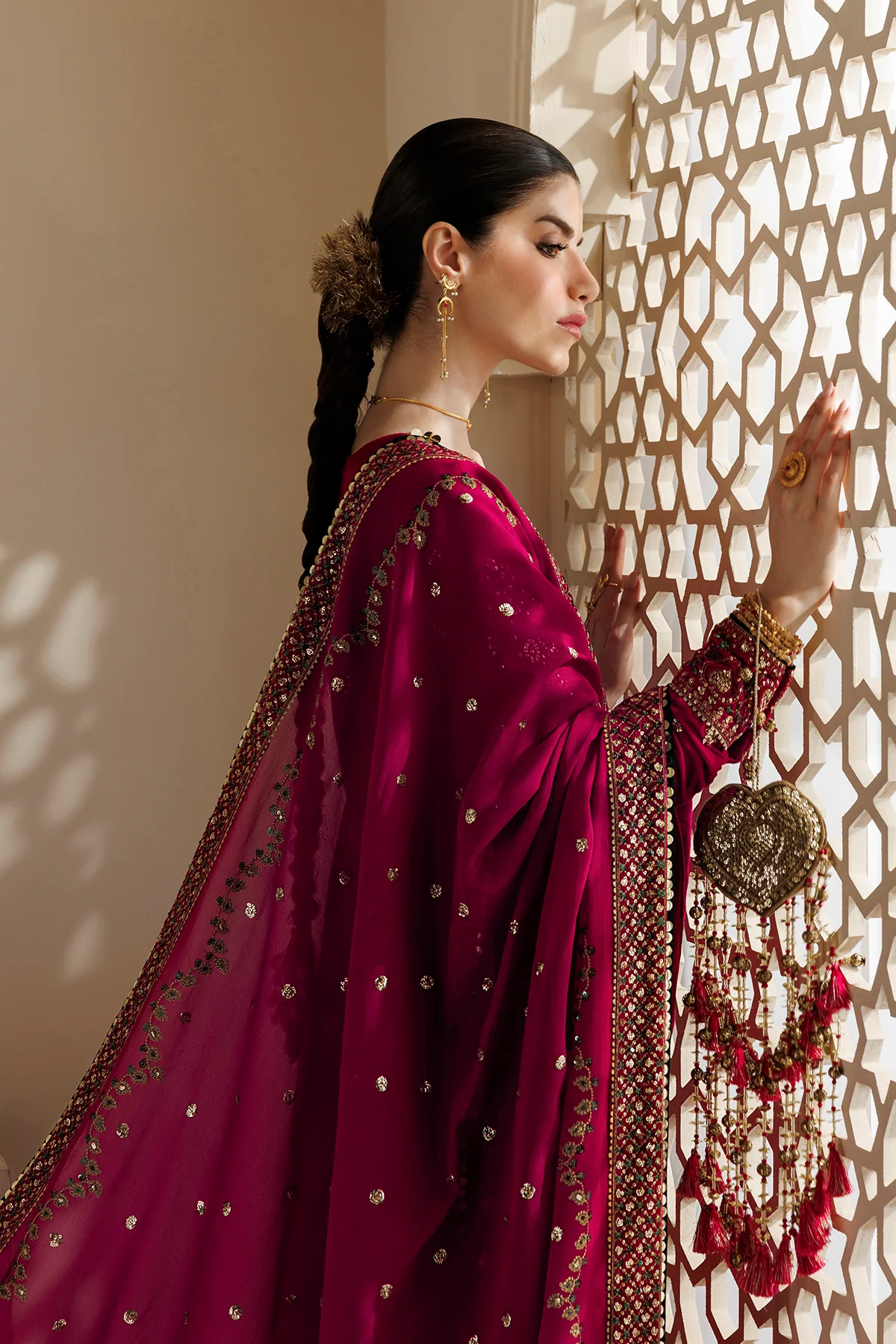 A woman in profile wearing a rich magenta-pink traditional outfit with gold embroidery, standing beside a decorative geometric screen that casts shadows on a beige wall.