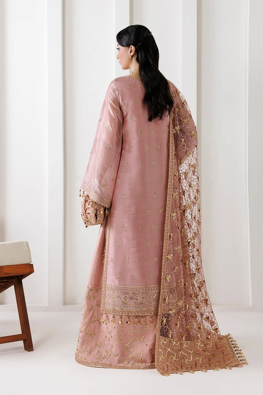 A woman shown from the back wearing a dusty pink traditional outfit with gold embroidery and a matching sheer dupatta draped over one shoulder, photographed in a bright indoor setting.