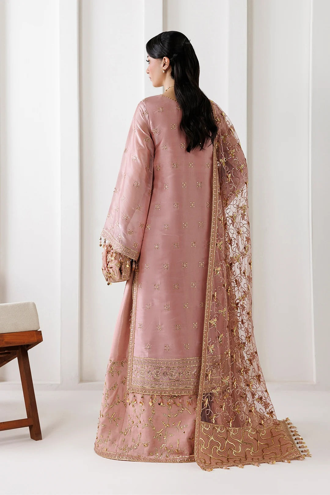 A woman shown from the back wearing a dusty pink traditional outfit with gold embroidery and a matching sheer dupatta draped over one shoulder, photographed in a bright indoor setting.