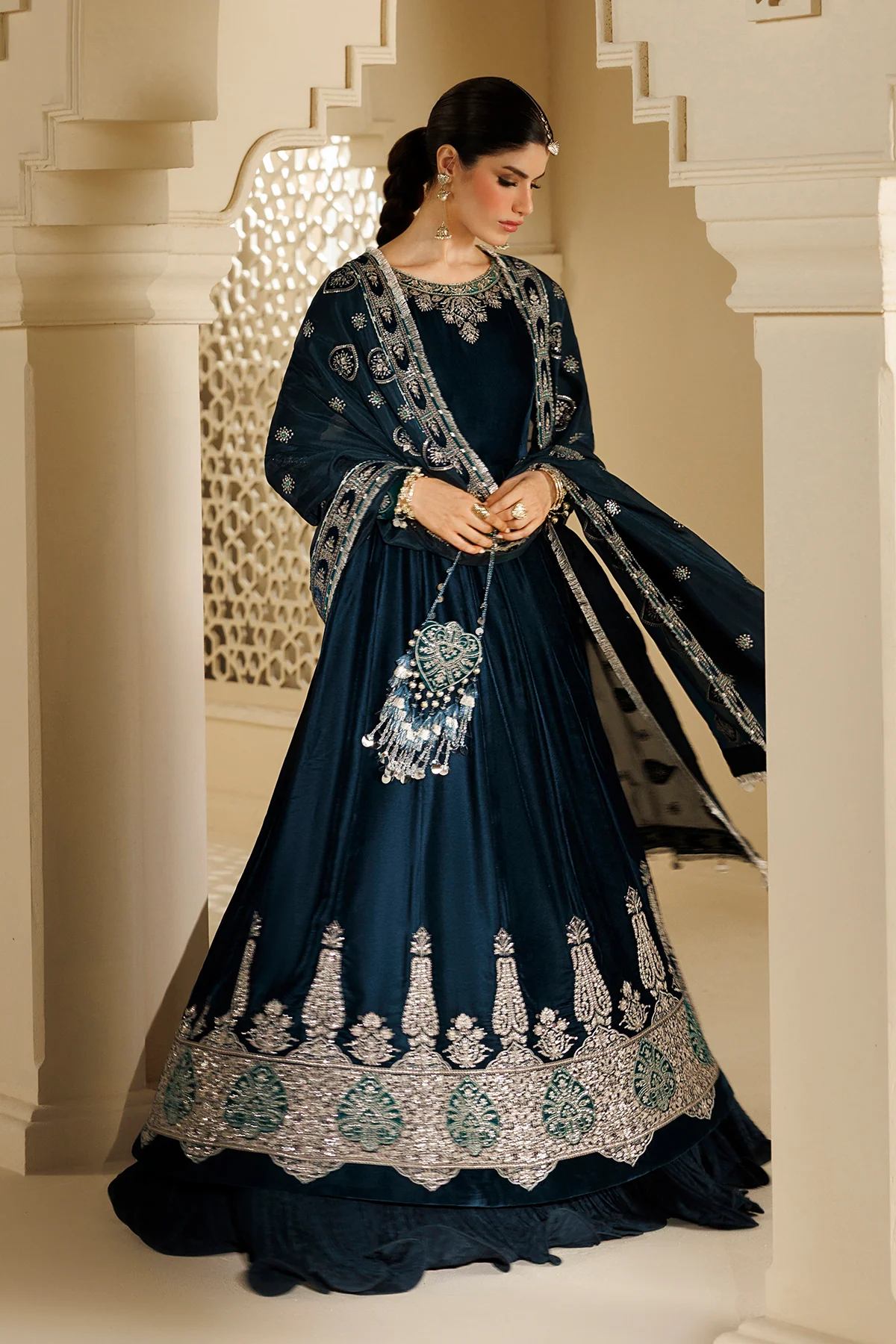 A woman models a dark navy blue traditional Indian anarkali gown with intricate silver and teal embroidery, paired with a matching dupatta and ornate jewelry, set against a classical architectural background with white pillars.