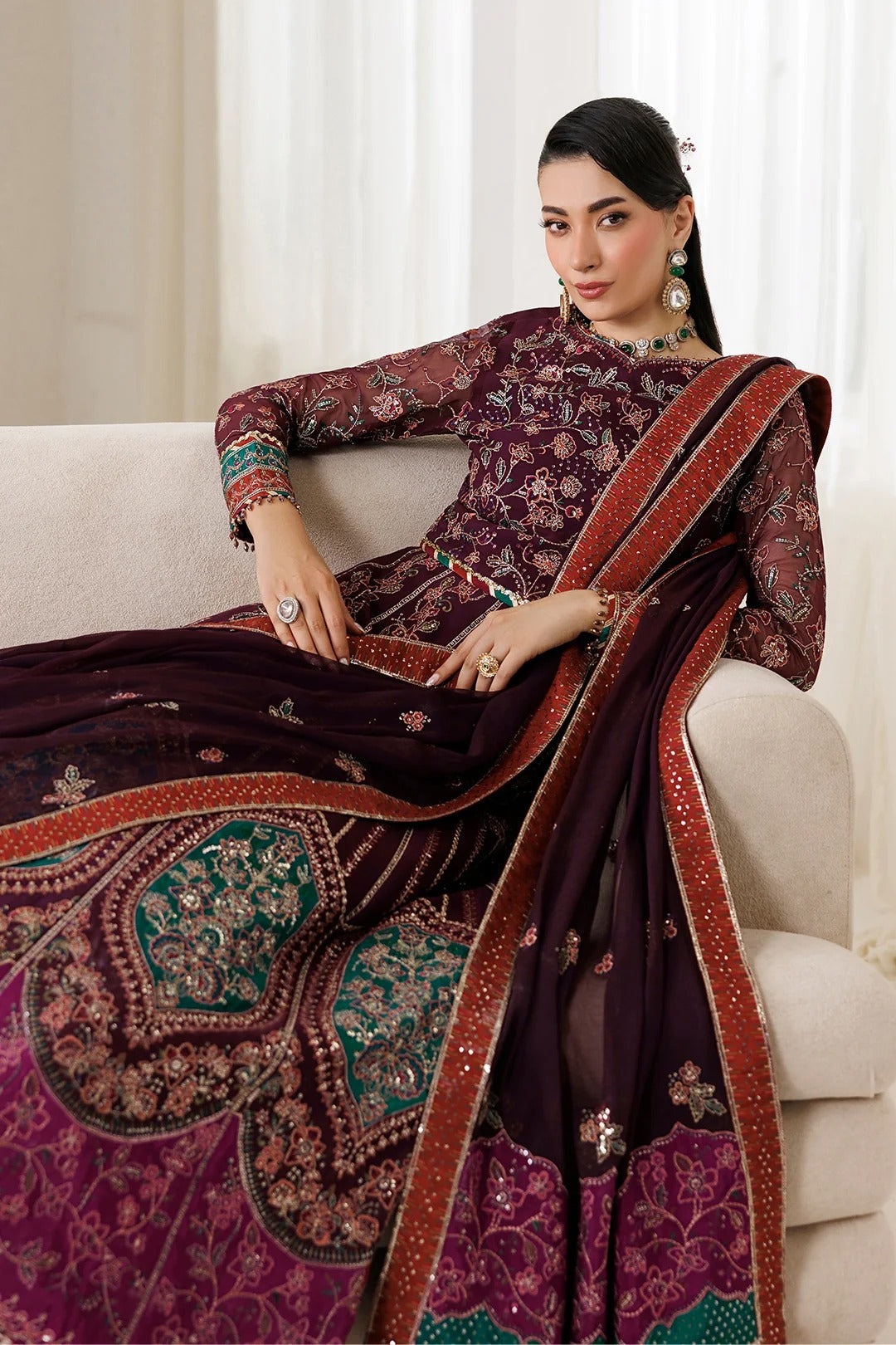 A woman models an ornate traditional Indian outfit consisting of a deep purple floral printed kurta and a matching dupatta draped over a light beige sofa. The dupatta features a wide red border with gold zari detailing and green and gold embroidered motifs.