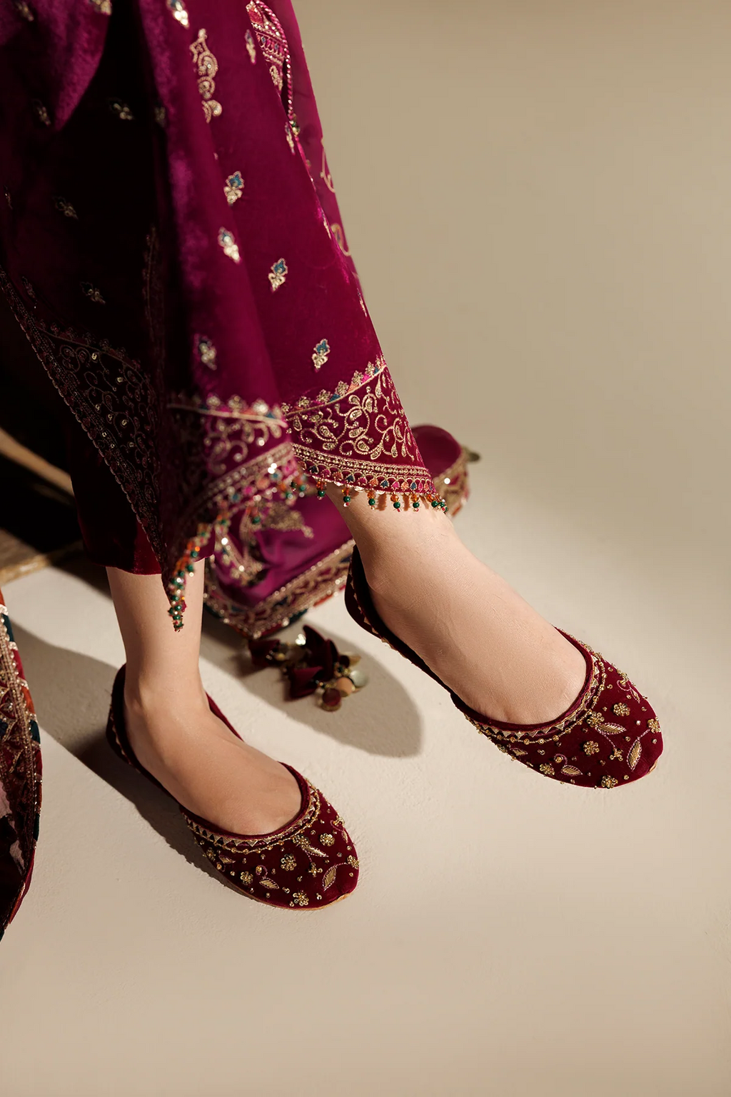 A close-up of a person wearing deep magenta embroidered flat shoes with traditional embellished pants, shown from the knees down against a plain beige studio background.