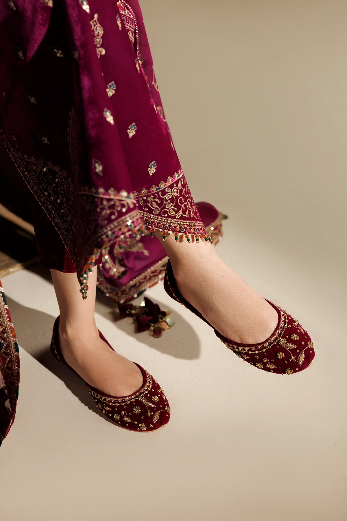 A close-up of a person wearing deep magenta embroidered flat shoes with traditional embellished pants, shown from the knees down against a plain beige studio background.