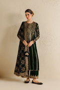 A woman models a dark green velvet traditional South Asian outfit with intricate gold embroidery, paired with matching flat shoes, standing against a textured beige wall.