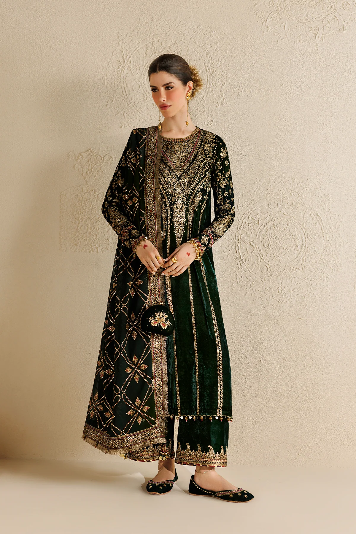 A woman models a dark green velvet traditional South Asian outfit with intricate gold embroidery, paired with matching flat shoes, standing against a textured beige wall.